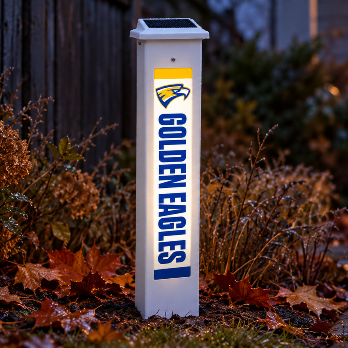 Wahlert Catholic Golden Eagles Solar Glow Post lit up at dusk in a garden bed of fallen leaves.